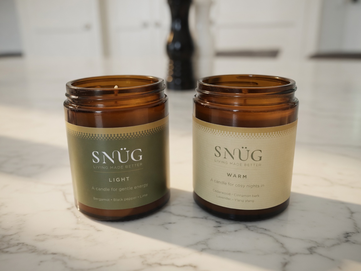 Two Snüg essential-oil soy candles, Light and Warm, in amber glass on a marble surface.