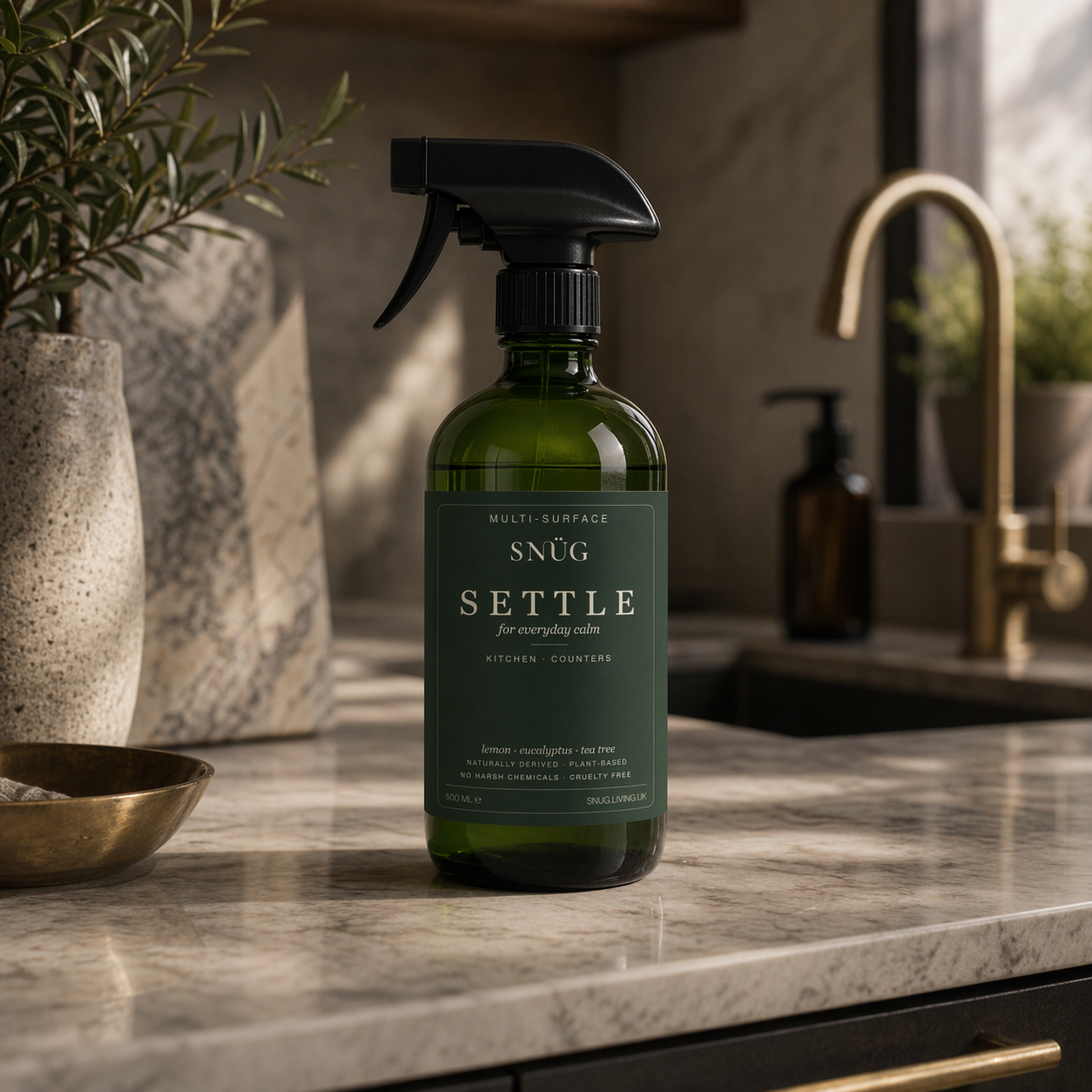 A Snüg Settle multi-surface spray in deep green glass, on a marble kitchen counter beside an olive sprig.
